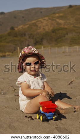 An infant girl in sunglasses lit by the late afternoon sun plays in the sand with a toy truck.