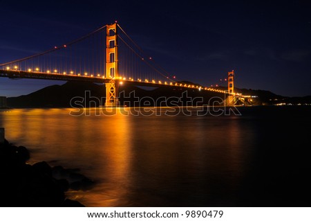 Golden Gate Bridge as seen from Fort Point overlook is glowing in the night with star trails in the sky behind it