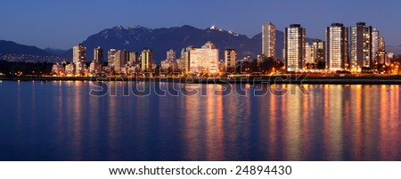 Highrises glow in gathering dusk on a winter Vancouver evening by the English Bay against a backdrop of majestic mountains.