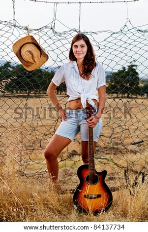 View of a beautiful young country girl with a guitar on the fence of the grassland.