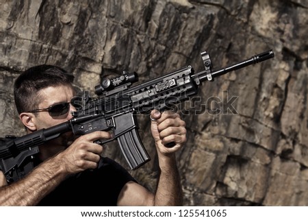 View of a menacing man pointing a machine gun in a black shirt and dark shades on a stone quarry.