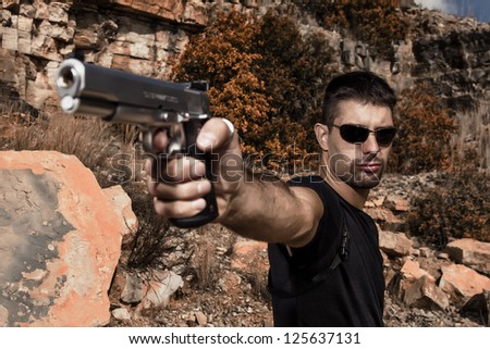 View of a menacing man pointing a handgun in a black shirt and dark shades on a stone quarry.