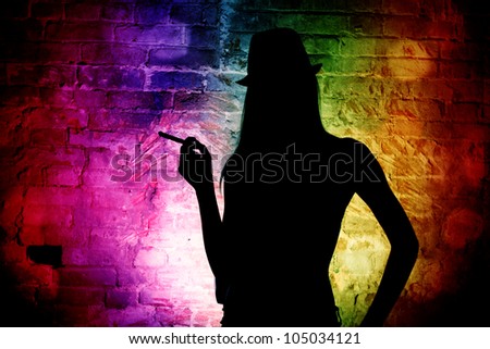 The dark silhouette of an elegant woman in a man\'s hat against a brick wall. Night club.