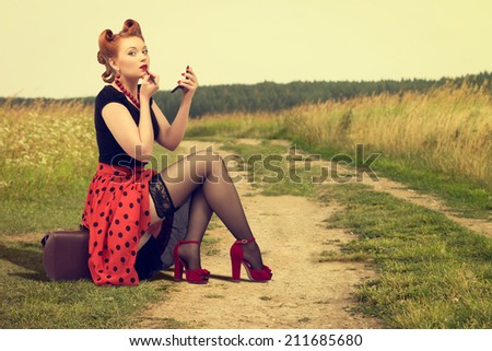Beautiful woman in pin up sitting on the side of a rural road painting lipstick.
