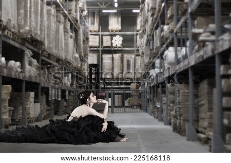 Beautiful young woman in a long black dress sitting in the middle of the production warehouse.