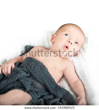 Baby with bright blue eyes looking up with a suprised look on his face laying on a white knit blanket with a gray knit blanket over him