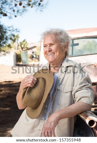 Smiling senior lady leaning against a vintage pickup truck holding a hat