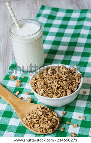 Almond breakfast granola with wood spoon on green checkered cloth with glass of milk vertical from above