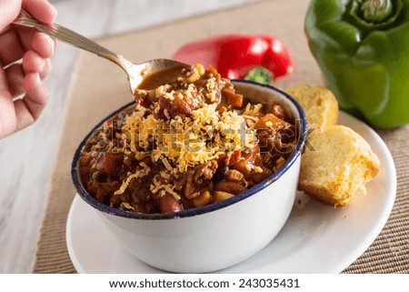 Bowl of warm chili winter comfort food dinner with corn bread muffin red and green peppers and hand scooping horizontal