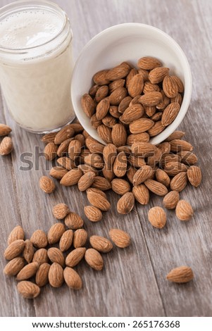 A shite bowl of almonds spilling onto wood background with glass of almond milk