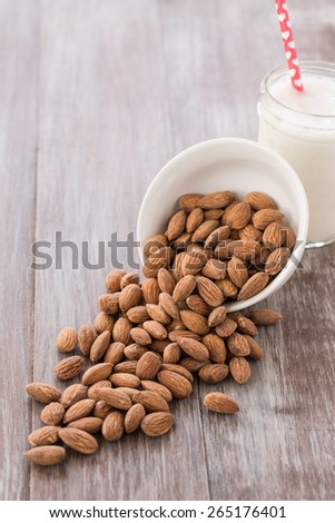 Almonds spilling out of white bowl onto wood background with glass of almond milk with heart straw in the background