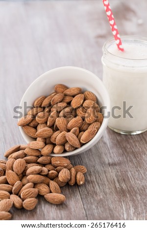 Almonds spilling out of white bowl onto wood background with glass of almond milk with heart straw