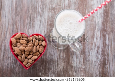 Almonds in a red heart shaped bowl with a glass of almond milk on wood background from above