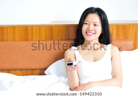Smiling attractive asian woman holding remote while changing channels on the television  in bedroom
