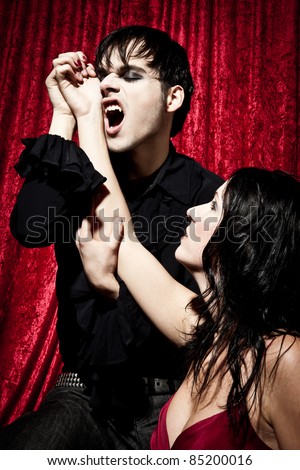 Male vampire is going to bite into a womans wrist. His mouth is open so you can see his fangs.