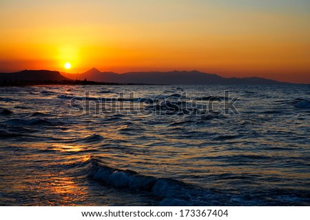 Fiery red sunset over a turbulent sea as the glowing orb of the sun sinks to the horizon over a distant mountain range in a spectacular marine sunset