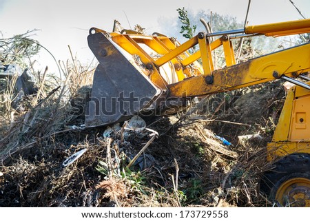 View of the bucket and hydraulic arm of an excavator clearing undergrowth and soil during construction work, while clearing a fire break