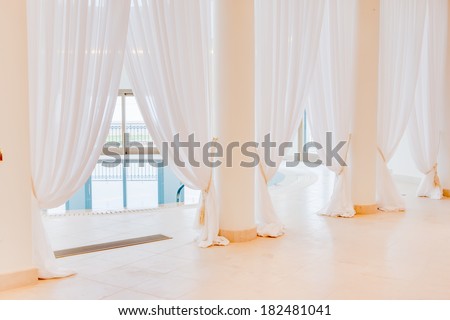 beautiful fresh clean floor length white drapes fastened to pillars in front of light airy windows surrounding an indoor swimming pool at a health resort or spa