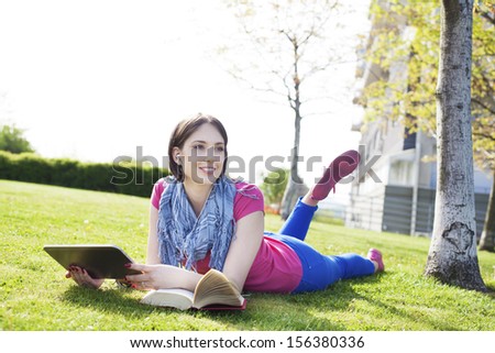 Beautiful happy young woman sitting on grass, enjoy sunny day and listening music with headphones and using digital tablet while reading book.