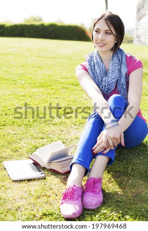 Beautiful happy young woman sitting on grass, enjoy sunny day and listening music with headphones and using digital tablet while reading book.