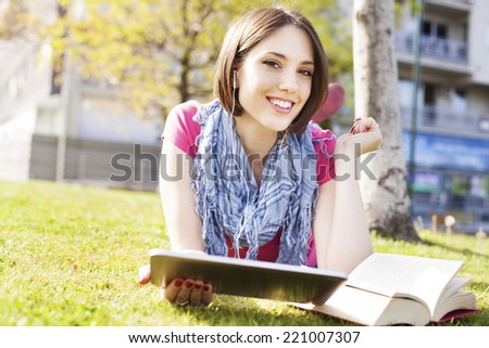 Beautiful happy young woman lying on grass, enjoy sunny day and listening music with headphones and using digital tablet while reading book.