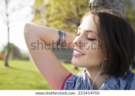 Portrait of a beautiful happy young woman sitting on grass, enjoy sunny day and listening music with headphones.