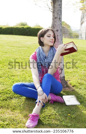 Beautiful happy young woman sitting on grass, enjoy sunny day and listening music with headphones while reading book, chilling.