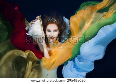 Red hair girl with big sea shell in colorful clothes on the dark background underwater