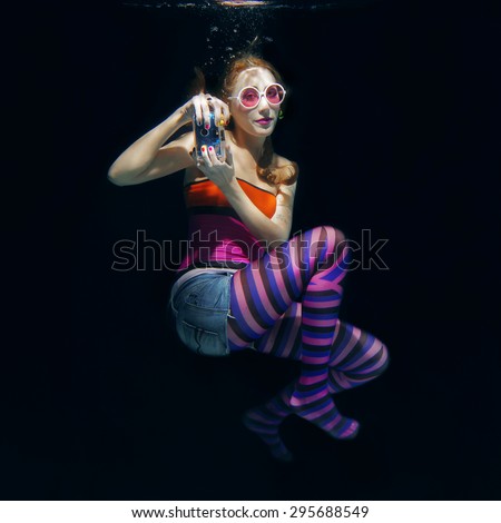 red hair funny girl in colorful clothes and pink sunglasses with tatto on the dark background swimming underwater