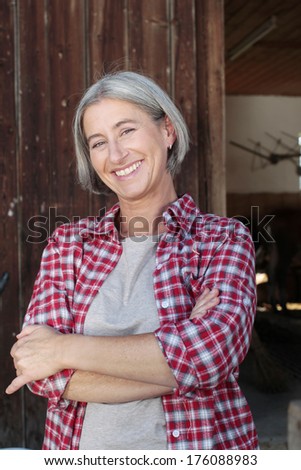 Matured farm woman with crossed arms in front of a barn smiling