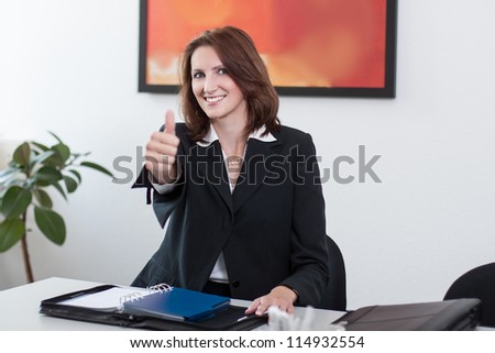 Young businesswoman sits in the office at the table and shows thumb up