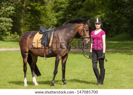 Girl stands besides her brown horse (New-Forest-Pony) on a meadow
