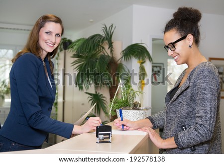 Smiling clerk (caucasian) is standing opposite to a customer (latin), who is filling out a document at a reception counter in the office.