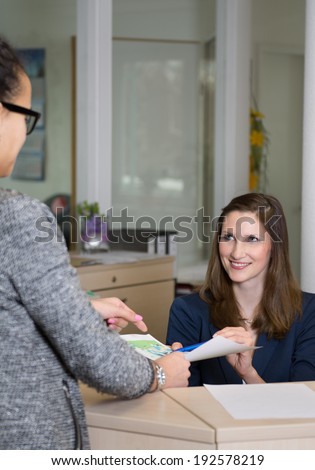 Smiling clerk (caucasian) is explaining a document to a client (latin) at a reception counter in the office. Woman in the background is in focus, woman in the foreground is blurred.