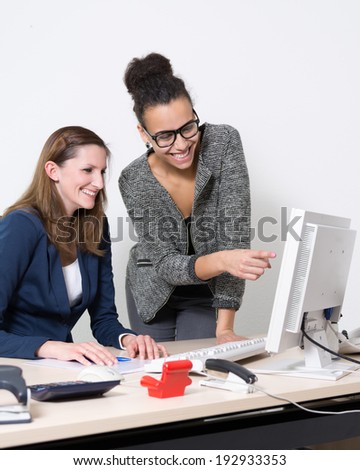 Two business women are sitting/standing in front of the computer in the office. Both women are smiling and looking to the monitor. One woman is pointing to the monitor.