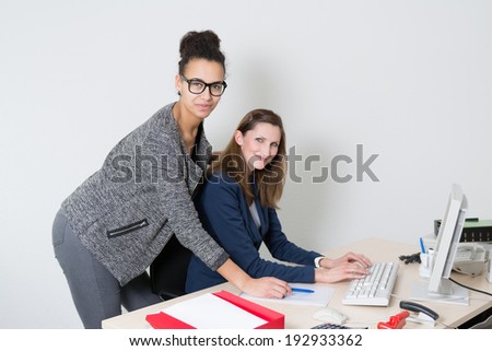 Two business woman are standing/sitting at the desk in the office. Both women are looking to the camera.