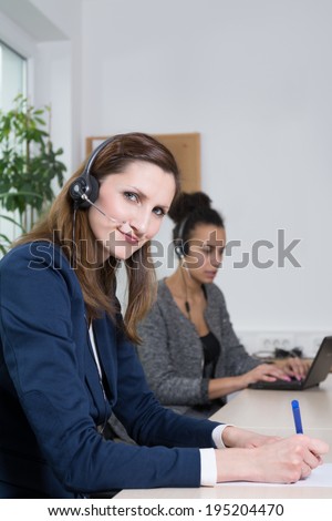 A young business woman with headset is writing at a document. Another woman with headset is sitting in the background (blurred) and is working at a notebook.