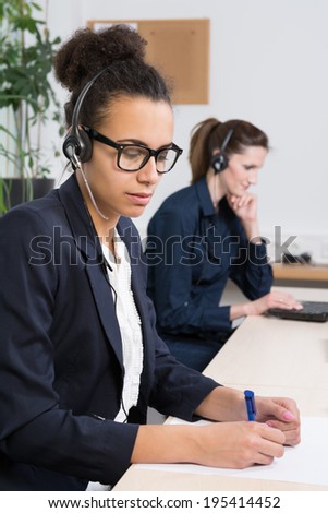 A young business woman with headset is writing at a document. Another woman with headset is sitting in the background (blurred) in front of a notebook.
