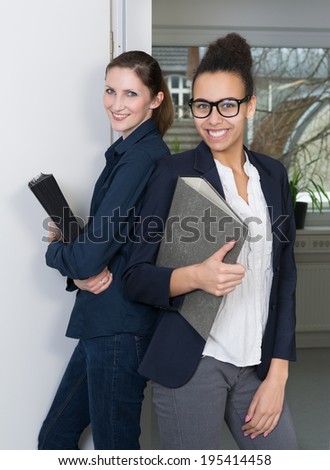 Two young business woman are standing side by side and are holding files in their hands.