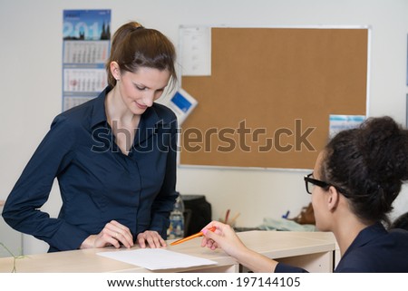 One woman is looking at a document which is lying at a counter in the office. Another woman sitting in front of the counter is pointing to the document with a pen.