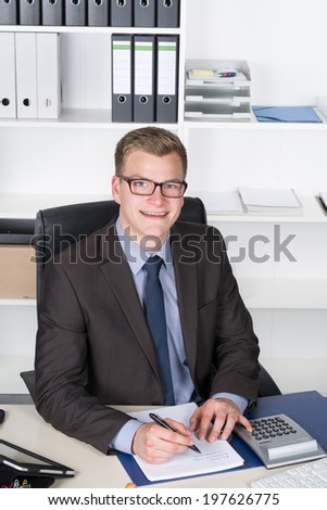 Young businessman is writing into a file while sitting at the desk in the office. The man is looking to the camera.