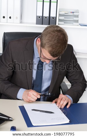 Young businessman with glasses is looking at a document through a magnifier while sitting at the desk in the office. A shelf is in the background.