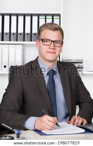 Young businessman is writing into a file while sitting at the desk in the office. A shelf is in the background. The man is looking to the camera.