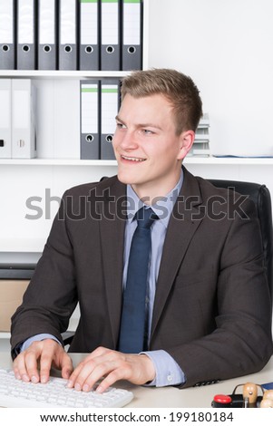 Young smiling businessman is typing at the computer keyboard while sitting at the desk in the office. A shelf is in the background. The man is looking sideways.