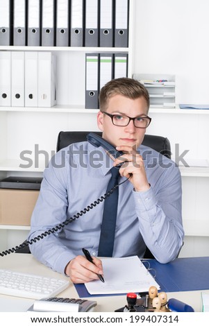 Young businessman with glasses is holding a telephone receiver and making notes in a file while sitting at the desk in the office. A shelf is in the background. The man is looking to the camera.