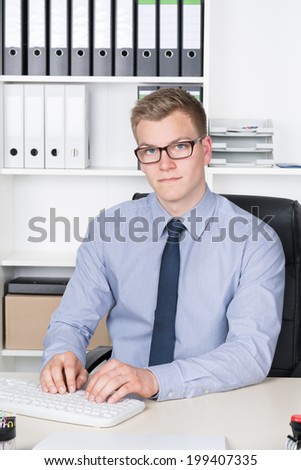 Young businessman is typing at the computer keyboard while sitting at the desk in the office. A shelf is in the background. The man is looking to the camera.