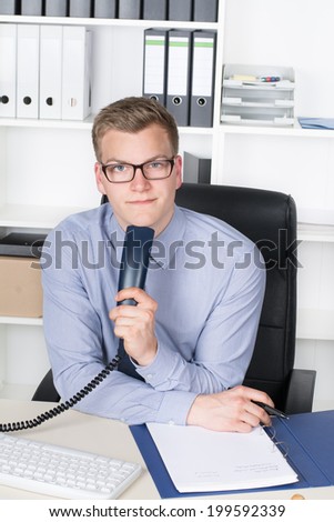 Young businessman with glasses is holding a telephone receiver and a pen while sitting at the desk in the office.