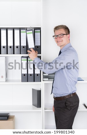 Young smiling businessman with glasses is taking out a file of a shelf in the office. The man is looking to the camera.