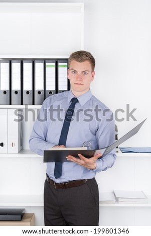 Young businessman is holding an opened file while standing in front of a shelf in the office. The man is looking to the camera.