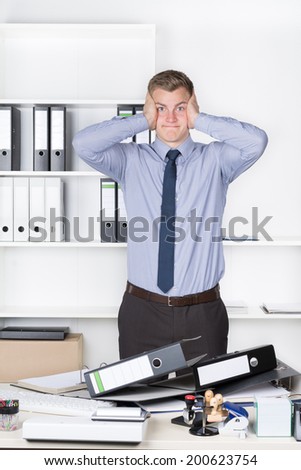 Young despaired businessman is standing in front of many files on his desk in the office. A shelf is in the background. The man is looking to the camera.